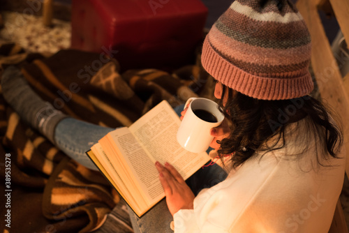 Young girl wearing a winter hat reading a book and having a cup of coffee on a cold winter night.