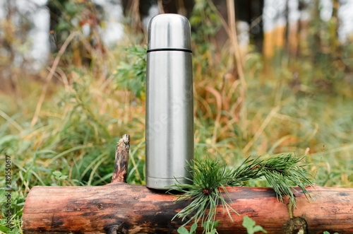 Thermos in a coniferous forest, close-up. Vacuum travel bottle stands on a tree stump. Concept for hiking, travel, hot drink in cold weather.