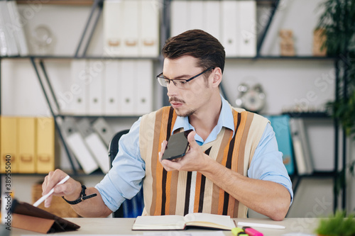 Fotografia Young entrepreneur talking to collegue on speakerphone when checking project on