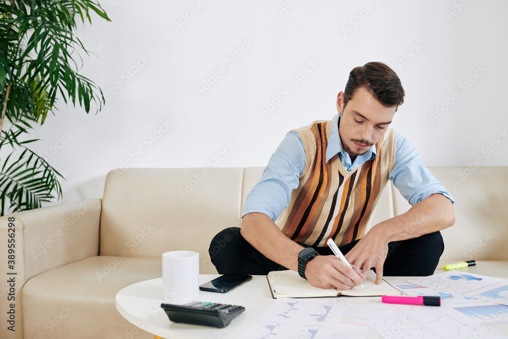Concentrated young businessman sitting at table with many financial documents and writing in planner Concentrated young businessman sitting at table with many financial documents and writing in planner