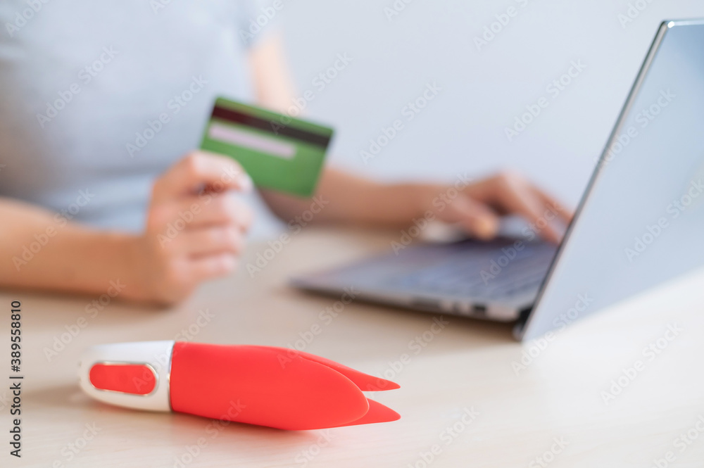 Fototapeta premium A faceless woman holds a credit card and a clitoral vibrator while sitting at a laptop. Online sex industry
