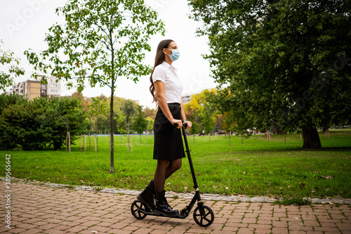 Businesswoman is riding push scooter in park. She is wearing protective face mask. Covid-19 virus pandemic concept.