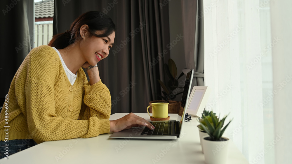 Side view of happy young woman looking on screen of laptop while sitting at home office. Side view of happy young woman looking on screen of laptop while sitting at home office.