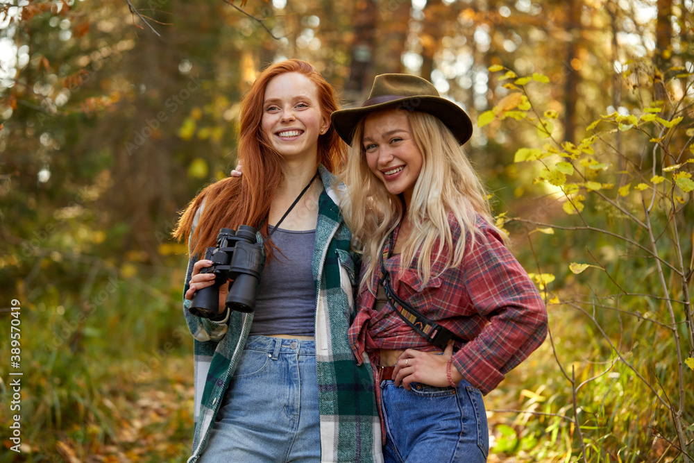 portrait of two young caucasian travellers women in the forest, inquisitive females enjoy hiking, nature, adventure, holidays. trekking, using binoculars, wearing clothes for hiking
