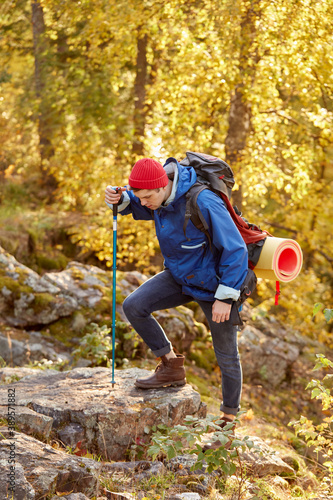 young caucasian male is concentrated on climbing mountains, holding bag on back and sportive wear, using stick for climbing, looking down on feet, in the nature