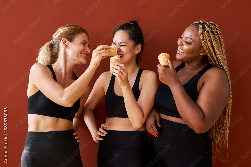 three diverse women eat ice-cream isolated on red background, interracial multi-ethnic team of women in black sportswear having fun, international friendship