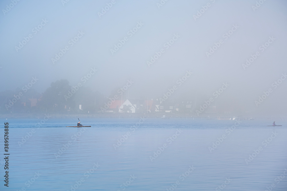 Stay fit rowing a canoe or kayak amidst the mist on a gloomy day. People keep fit kayaking amid tranquility on a still, calm Baltic sea as the coast is visible through the fog - Dragor Havn, Denmark