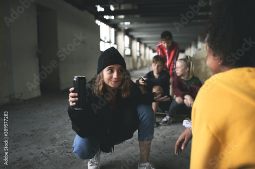 Group of teenagers gang sitting indoors in abandoned building, using smartphones.