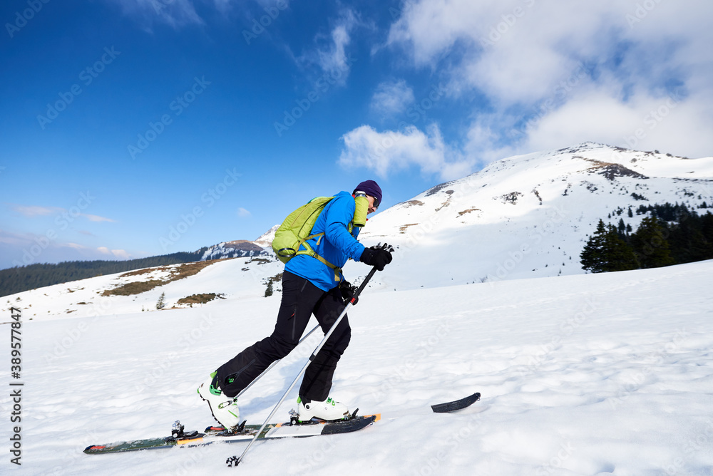 Happy tourist in colorful clothing and sunglasses with backpack climbing on skis in deep snow on background of bright blue sky and beautiful mountain. Winter vacations, active lifestyle concept.