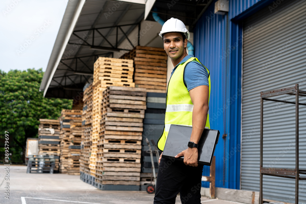 Fototapeta premium Portrait of young Indian worker working in logistic industry outdoor in front of factory warehouse. Smiling happy man with hard hat looking at camera walking and holding a laptop at depot