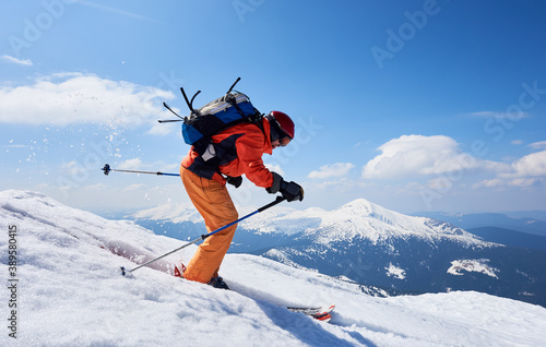 Sportsman skier in skiing equipment with backpack riding down steep slope in deep snow on copy space background of blue sky and beautiful mountain landscape. Winter sports, courage and speed concept.