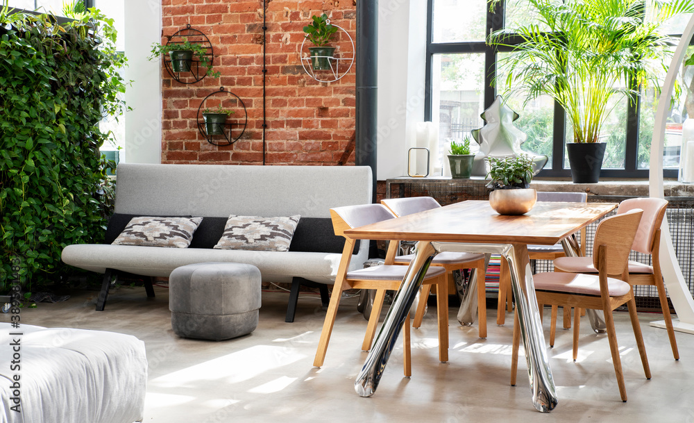 Interior of industrial room in loft apartment with vertical garden, wooden stylish table and chairs, big window, sofa and brick wall. Living room with dining room. Banner
