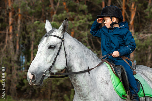 Little boy in a jockey cap on a white adult horse on a background of nature. Jockey, hippodrome, horseback riding © Aliaksandr Marko