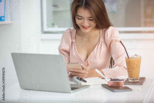Image of an Asian business woman working on a computer, a phone and a laptop with books and coffee cups on a white table in the morning sitting by the window. At her office