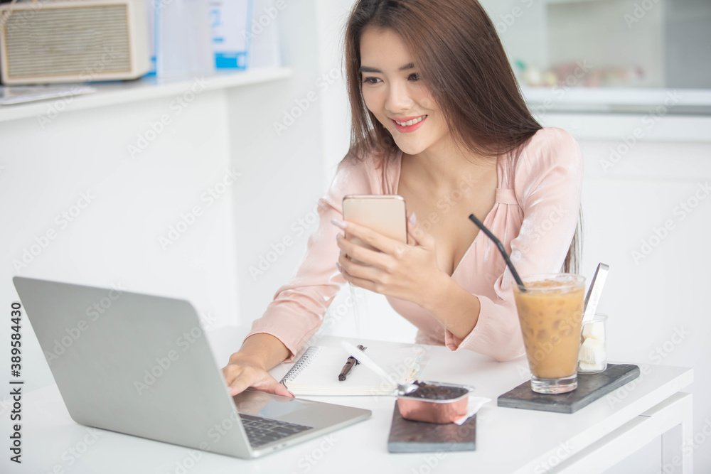 Image of an Asian business woman working on a computer, a phone and a laptop with books and coffee cups on a white table in the morning sitting by the window. At her office Image of an Asian business woman working on a computer, a phone and a laptop with books and coffee cups on a white table in the morning sitting by the window. At her office