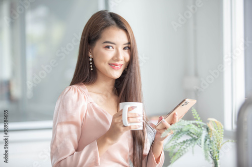 Image of an Asian business woman working on a computer, a phone and a laptop with books and coffee cups on a white table in the morning sitting by the window. At her office