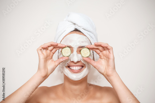 Funny smiling Spa Woman with towel wrapped around her head, applying fresh Facial Mask with cucumbers. Beauty Treatments. Face mask, skin care concept