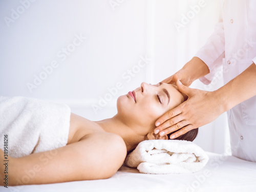 Beautiful brunette woman enjoying facial massage with closed eyes in sunny spa center . Relaxing treatment and cosmetic medicine concepts