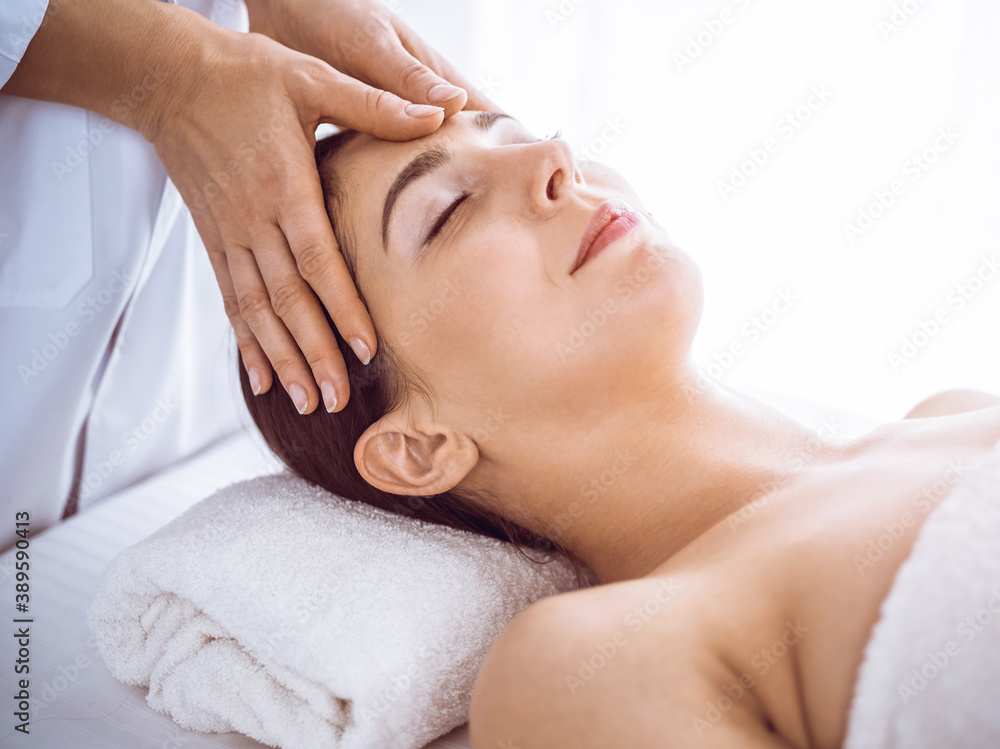 Beautiful brunette woman enjoying facial massage with closed eyes in sunny spa center . Relaxing treatment and cosmetic medicine concepts