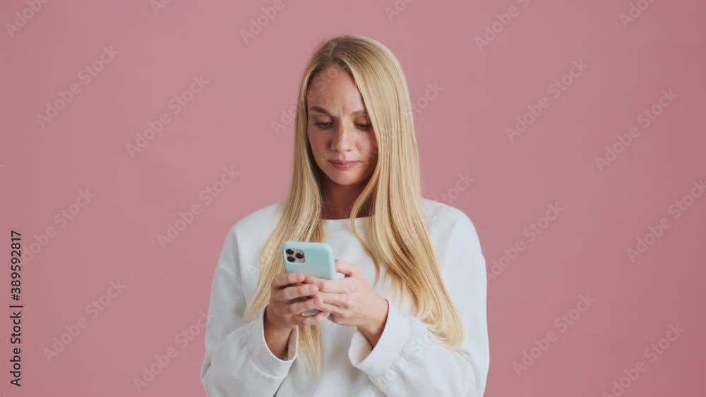 An attractive blonde woman is looking to the side while using her smartphone standing isolated over pink background in studio An attractive blonde woman is looking to the side while using her smartphone standing isolated over pink background in studio