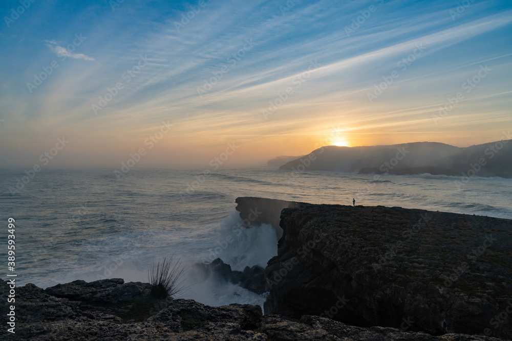 huge storm surge ocean waves crashing onto shore and cliffs at sunrise with a person standing there