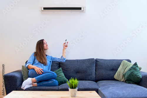 Valokuva Young happy woman sitting on couch under air conditioner and adjusting comfort t