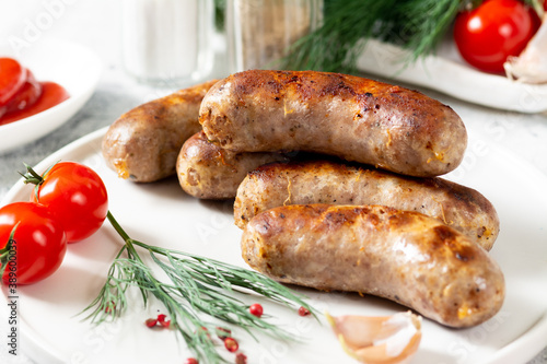 Fried sausages. Fried sausages with spices, sauce, tomatoes and parsley. Delicious meat sausages in a white ceramic plate on a light gray table. German dish	