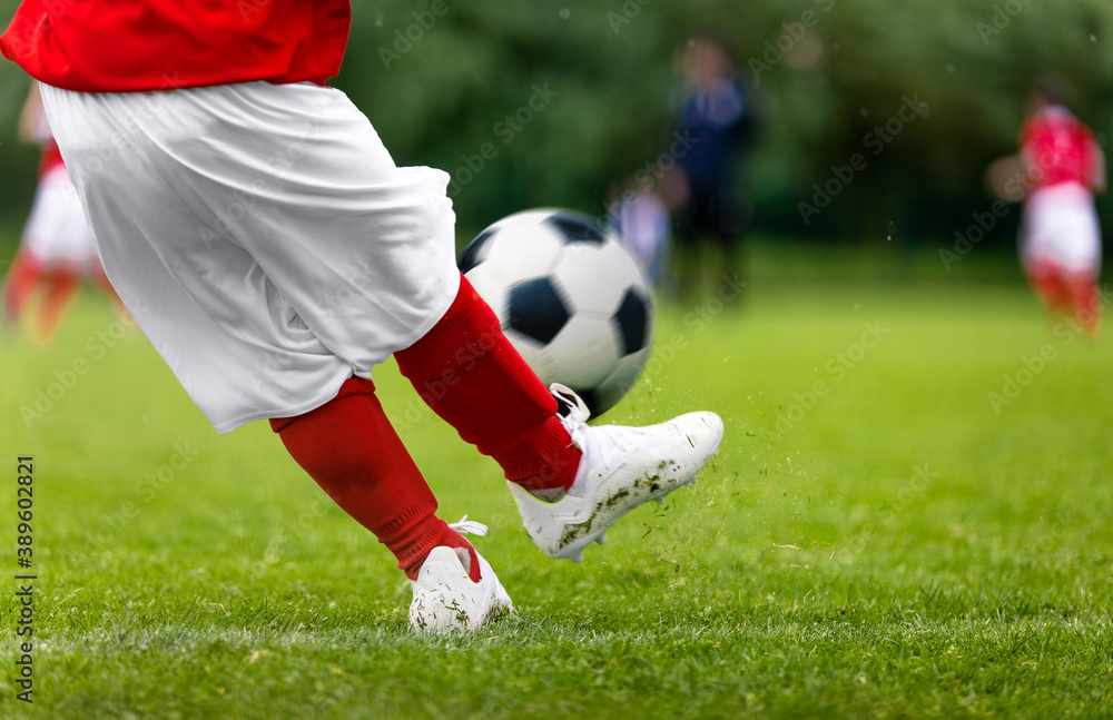 Football kick. Young player kicking soccer ball on green grass turf field. Closeup of footballers feet in cleats playing soccer ball. Red and white sports clothing. Junior level soccer sportswear Football kick. Young player kicking soccer ball on green grass turf field. Closeup of footballers feet in cleats playing soccer ball. Red and white sports clothing. Junior level soccer sportswear