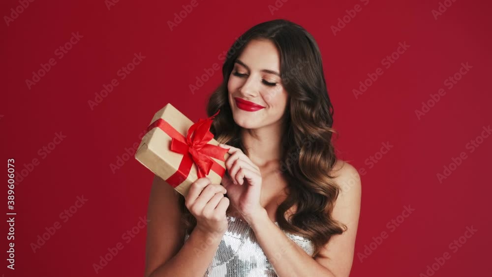 A smiling woman is holding a present standing isolated over a red background in the studio A smiling woman is holding a present standing isolated over a red background in the studio