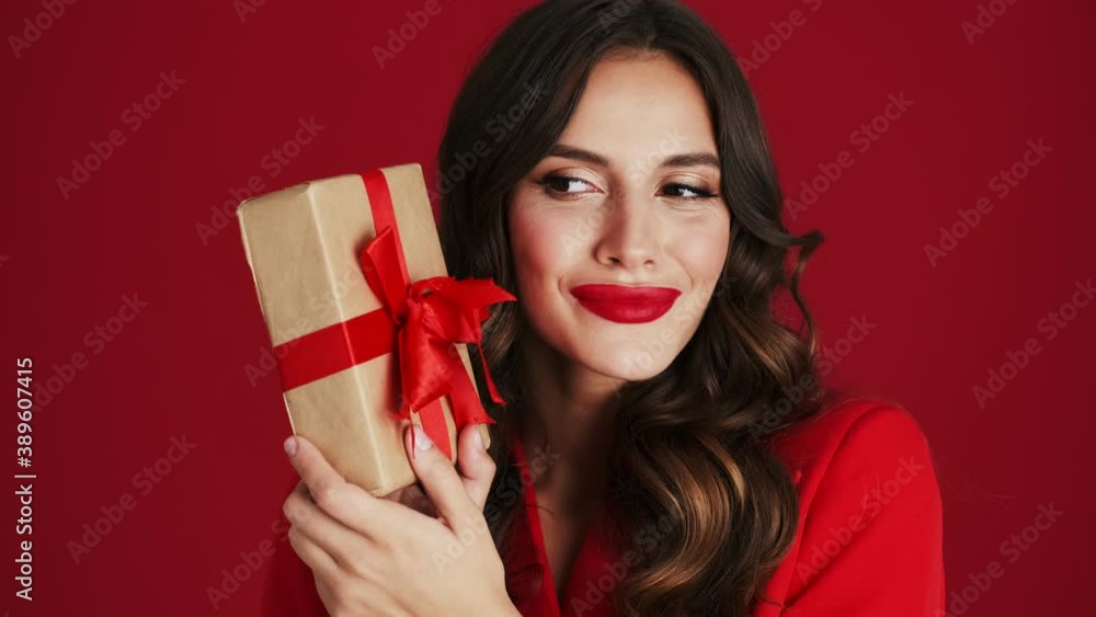 Close-up view of a cheerful woman in a red blazer is shaking a gift standing isolated over red background in the studio Close-up view of a cheerful woman in a red blazer is shaking a gift standing isolated over red background in the studio