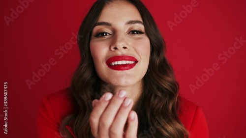 Close-up view of a smiling woman in a red blazer is sending an air kiss standing isolated over red background in the studio photo