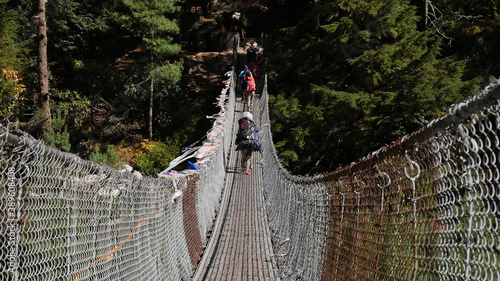 Porters crossing a suspension bridge with tied Buddhist prayer flags spanning Dudhkoshi River in a valley near Manjo on Mount Everest Base Camp Trek, Nepal. photo