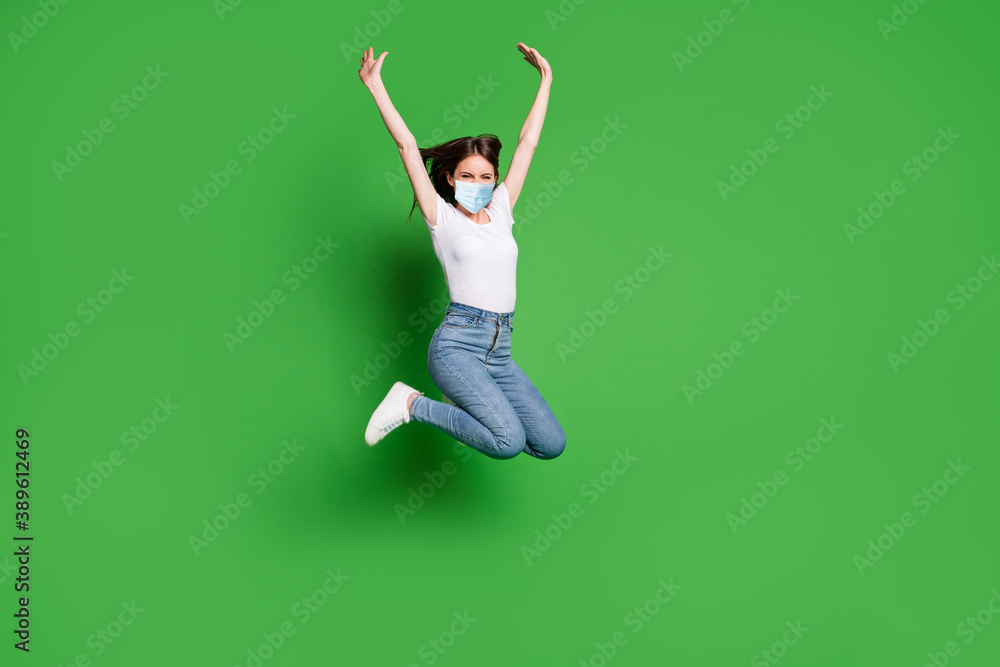 Full size photo of careless girl jump raise arms wear medical safety mask casual style clothes isolated green color background Full size photo of careless girl jump raise arms wear medical safety mask casual style clothes isolated green color background