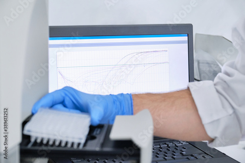 A researcher putting PCR plate on the thermal cycler for DNA amplification. Curve chart on monitor behind on focus. Coronavirus PCR test. photo