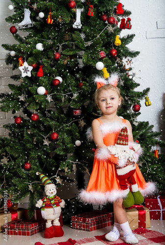 Christmas, New Year tree. A little girl in a fluffy, red dress and ears on her head is standing under the Christmas tree.