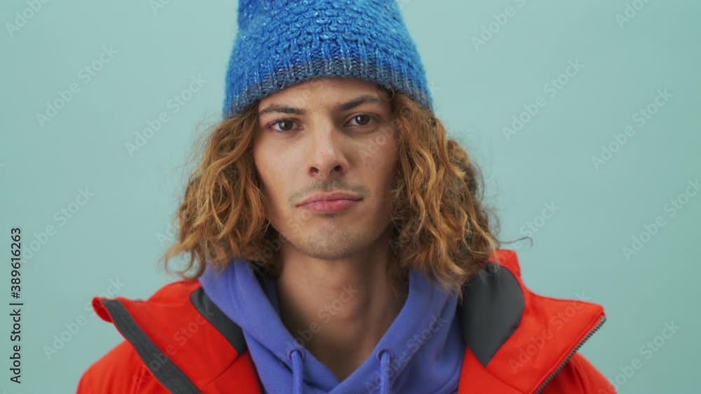 A handsome snowboarder is winking while looking to the camera standing isolated over a blue background in the studio A handsome snowboarder is winking while looking to the camera standing isolated over a blue background in the studio