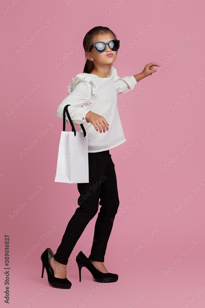 fashionable adorable little child girl in sunglasses and oversized mother's shoes is holding shopping bags on pink background. fashionable adorable little child girl in sunglasses and oversized mother's shoes is holding shopping bags on pink background.