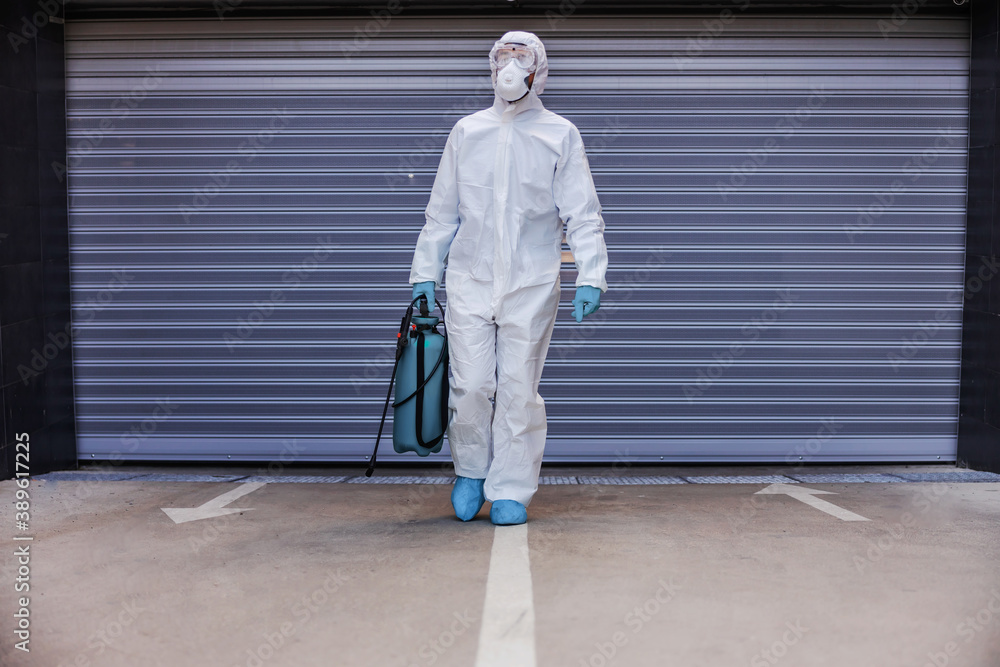 Man in white sterile uniform with rubber gloves holding sprayer with disinfectant and walking towards camera in garage.