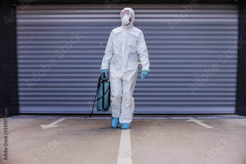 Man in white sterile uniform with rubber gloves holding sprayer with disinfectant and walking towards camera in garage.