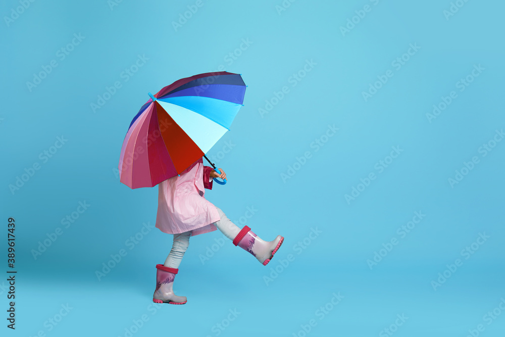 funny little child girl with multicolored umbrella in pink rain coat and rubber boots on blue background. copy space funny little child girl with multicolored umbrella in pink rain coat and rubber boots on blue background. copy space
