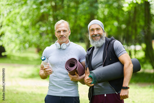 Medium portrait shot of two joyful senior sportsmen spending time in park holding yoga mats looking at camera