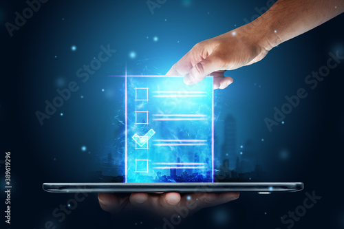 Online voting, Hand with a hologram ballot and a box for Internet voting in a mobile phone on a blue background. Mixed environment, e-voting technology concept. photo