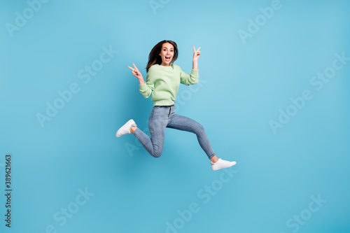 Full length body size photo of funky friendly girl showing v-sign gesture with both hands jumping high smiling isolated on vivid blue color background