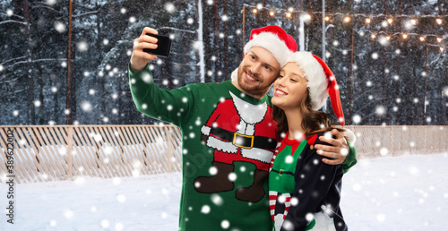 people, technology and holidays concept - happy couple in santa hats and ugly sweaters taking selfie with smartphone over skating rink and snow on background