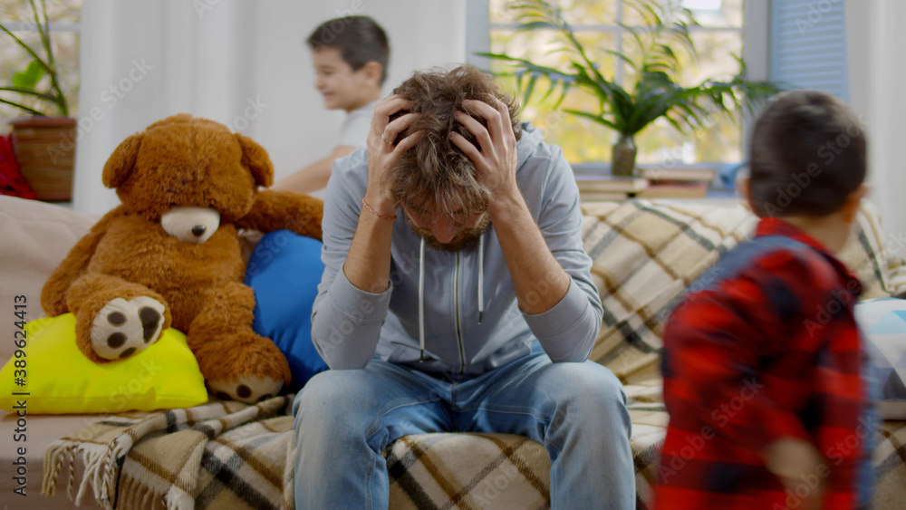 Young man sitting on couch tired of noisy children running and shouting Young man sitting on couch tired of noisy children running and shouting