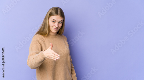Young blonde woman isolated on purple background stretching hand at camera in greeting gesture.