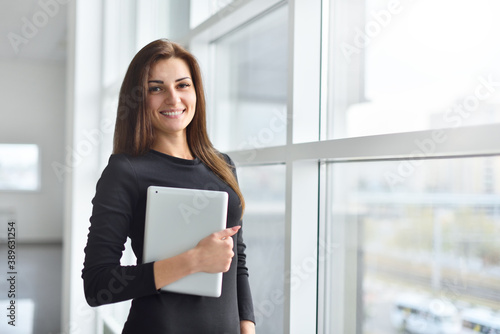 Young woman office manager stands by the window and holds a digital tablet in her hands