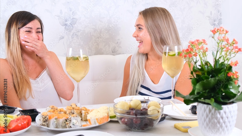 Two attractive girls, cheerful best friends having fun and drinking wine at home. Two glasses of white wine in hands. Girls having fun time together gossiping. Two attractive girls, cheerful best friends having fun and drinking wine at home. Two glasses of white wine in hands. Girls having fun time together gossiping.
