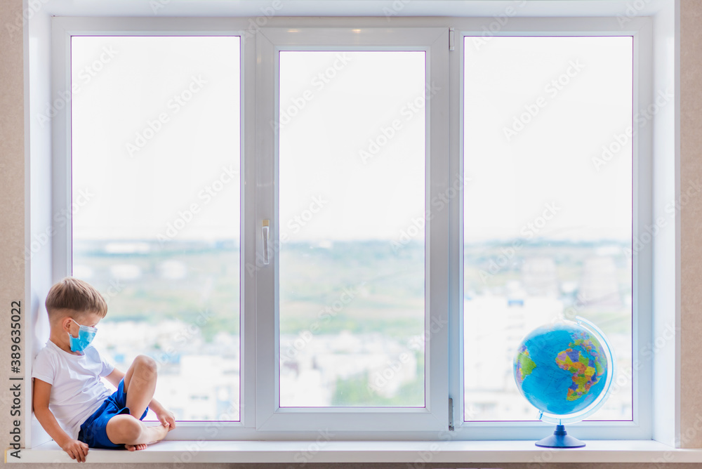 The masked kid sits at home in quarantine. The child is examining the globe while sitting on the windowsill. Prevention of coronavirus Covid-19 during a pandemic. The masked kid sits at home in quarantine. The child is examining the globe while sitting on the windowsill. Prevention of coronavirus Covid-19 during a pandemic.