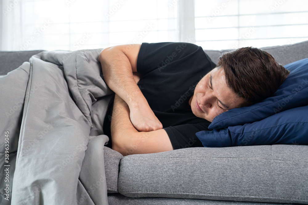 Sick and unwell Asian men holding his head, having a strong headache and sleeping on sofa at home wrapped in grey blanket Sick and unwell Asian men holding his head, having a strong headache and sleeping on sofa at home wrapped in grey blanket